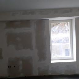 GmbH Krzysztof Fischer - Ściana w trakcie przygotowywania do malowania, z widocznymi łatami i szpachlą, okno zabezpieczone folią ochronną, widoczne gniazdka elektryczne.