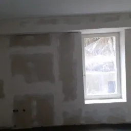 Ściana w trakcie przygotowywania do malowania, z widocznymi łatami i szpachlą, okno zabezpieczone folią ochronną, widoczne gniazdka elektryczne.