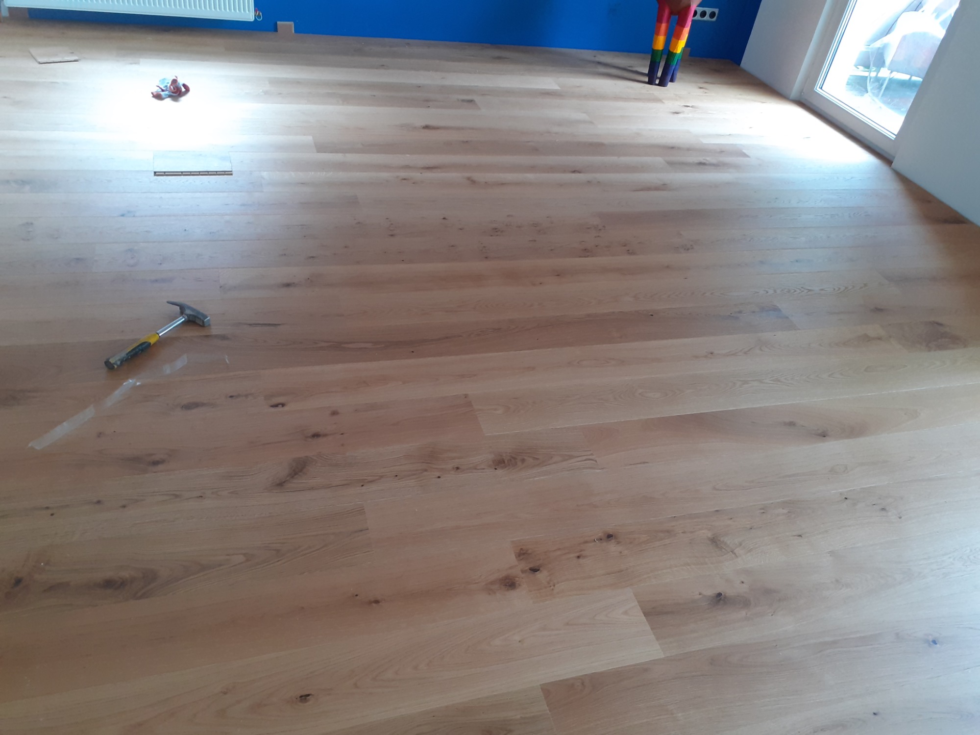 Jasna, drewniana podłoga ułożona z paneli, widoczny młotek z żółtą rękojeścią, fragment niebieskiej ściany i grzejnika w tle, w rogu pokoju tęczowe nogi od krzesła lub stołu.