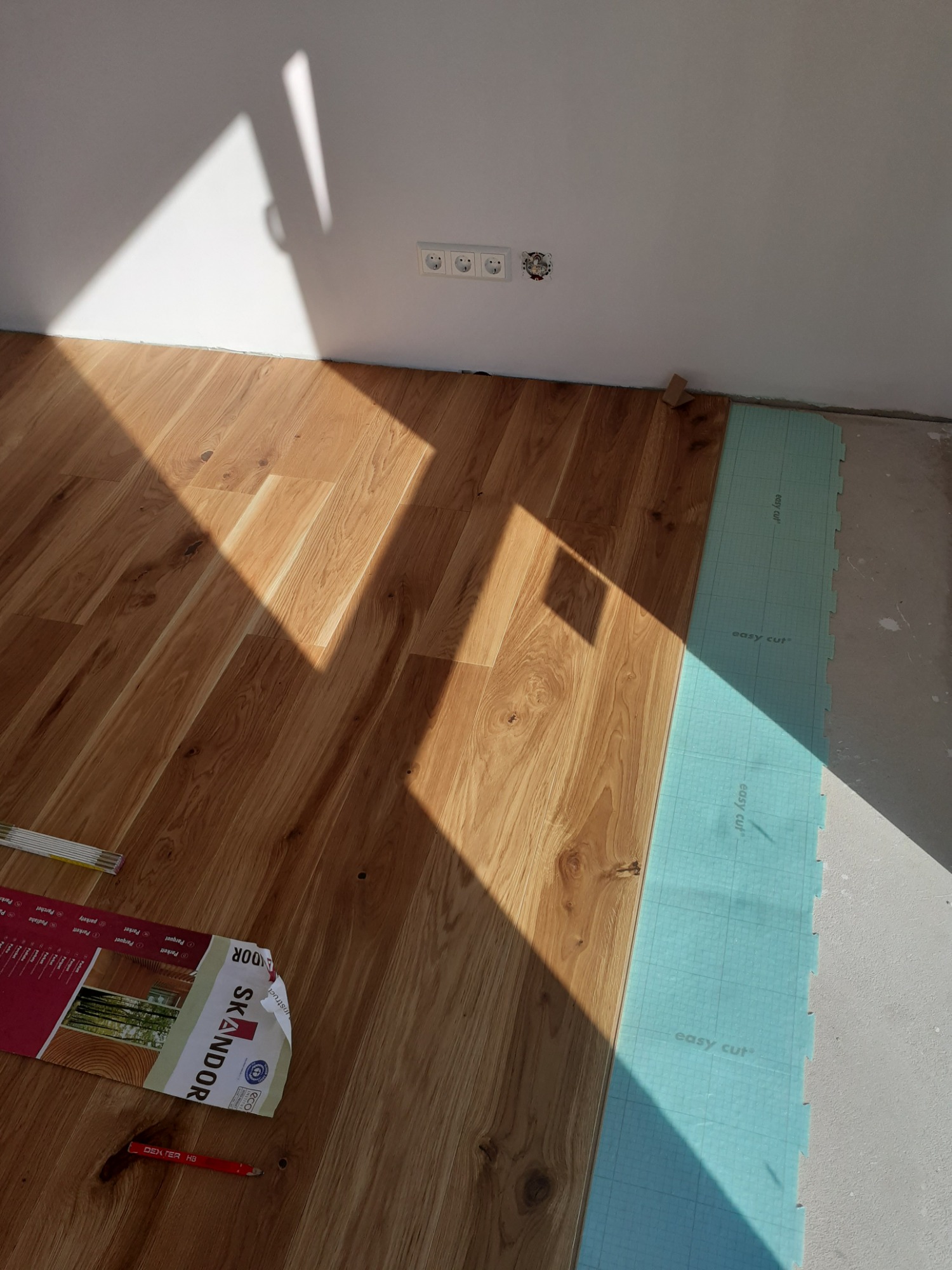 Układanie drewnianej podłogi: widok na panele, podkład wyciszający 'easy cut', paczkę paneli SKANDOR, ołówek i miarkę w słonecznym wnętrzu.