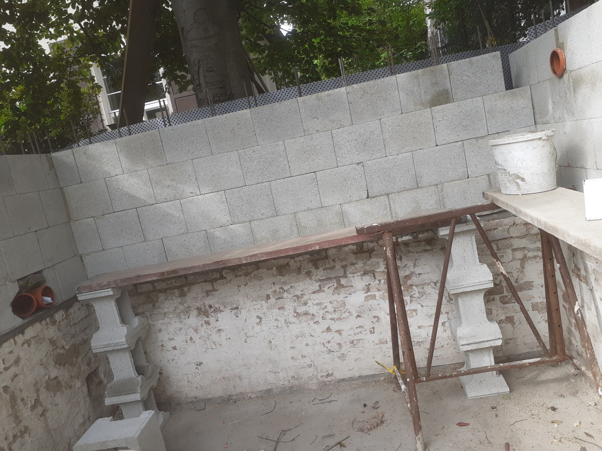 Wznoszenie muru z szarego bloczka betonowego, widoczne szalunki, podpory z prefabrykatów betonowych, fragment starego muru z cegły, rury kanalizacyjne w ścianie, wiaderko na rusztowaniu.