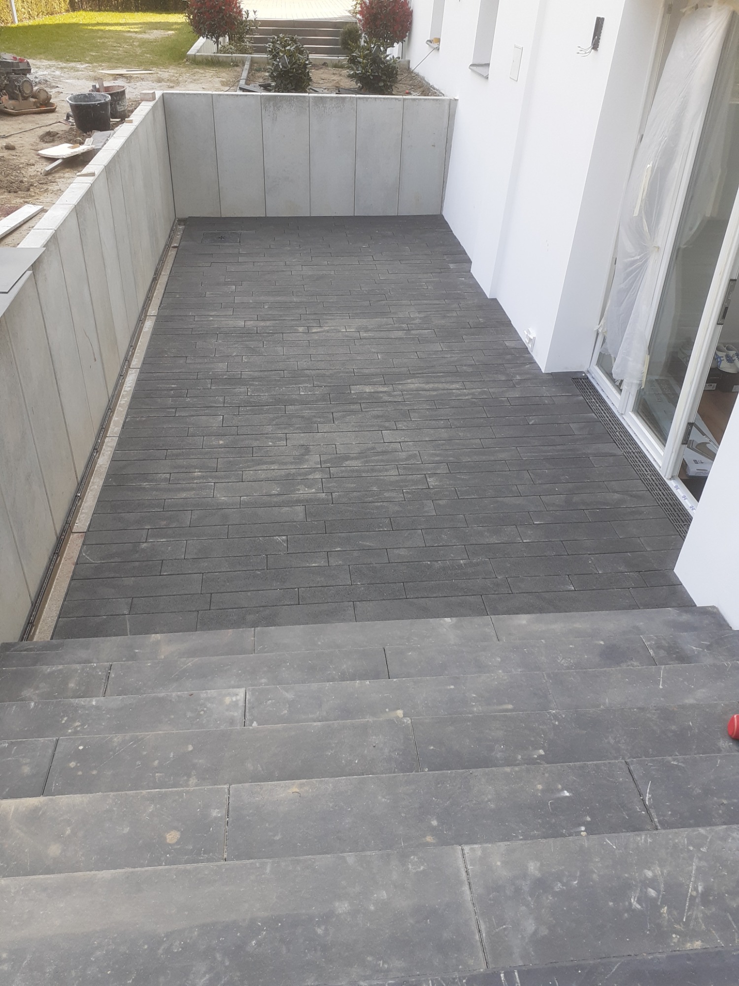 Ułożona kostka brukowa na patio z niskimi schodami, widoczny odpływ liniowy przy ścianie budynku.