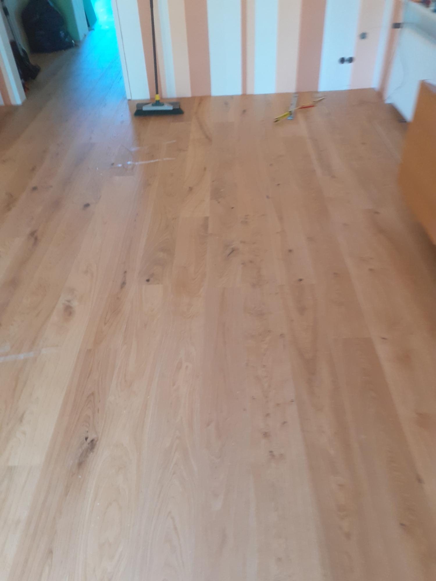 Jasna drewniana podłoga po remoncie, widoczna szczotka do zamiatania i poziomica z ołówkiem przy ścianie w pasy.