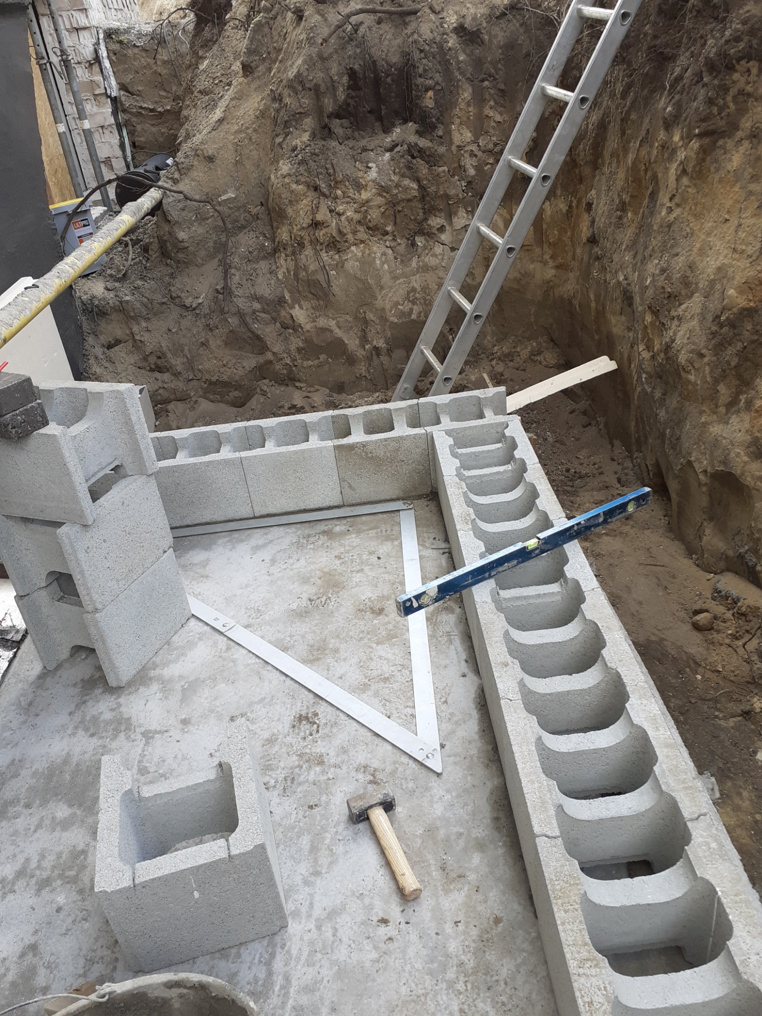 Fundamenty z bloczków betonowych szalunkowych układane w wykopie, z użyciem poziomicy, młotka i drabiny opierającej się o ścianę wykopu.