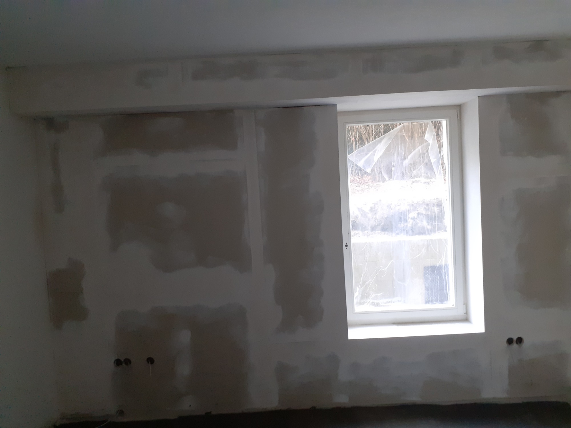 Ściana w trakcie przygotowywania do malowania, z widocznymi łatami i szpachlą, okno zabezpieczone folią ochronną, widoczne gniazdka elektryczne.