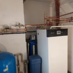 Instalacja grzewcza z białą pompą ciepła Heiztechnik, miedzianymi rurami, niebieskim zbiornikiem wyrównawczym i filtrem wody Multitower w kotłowni.