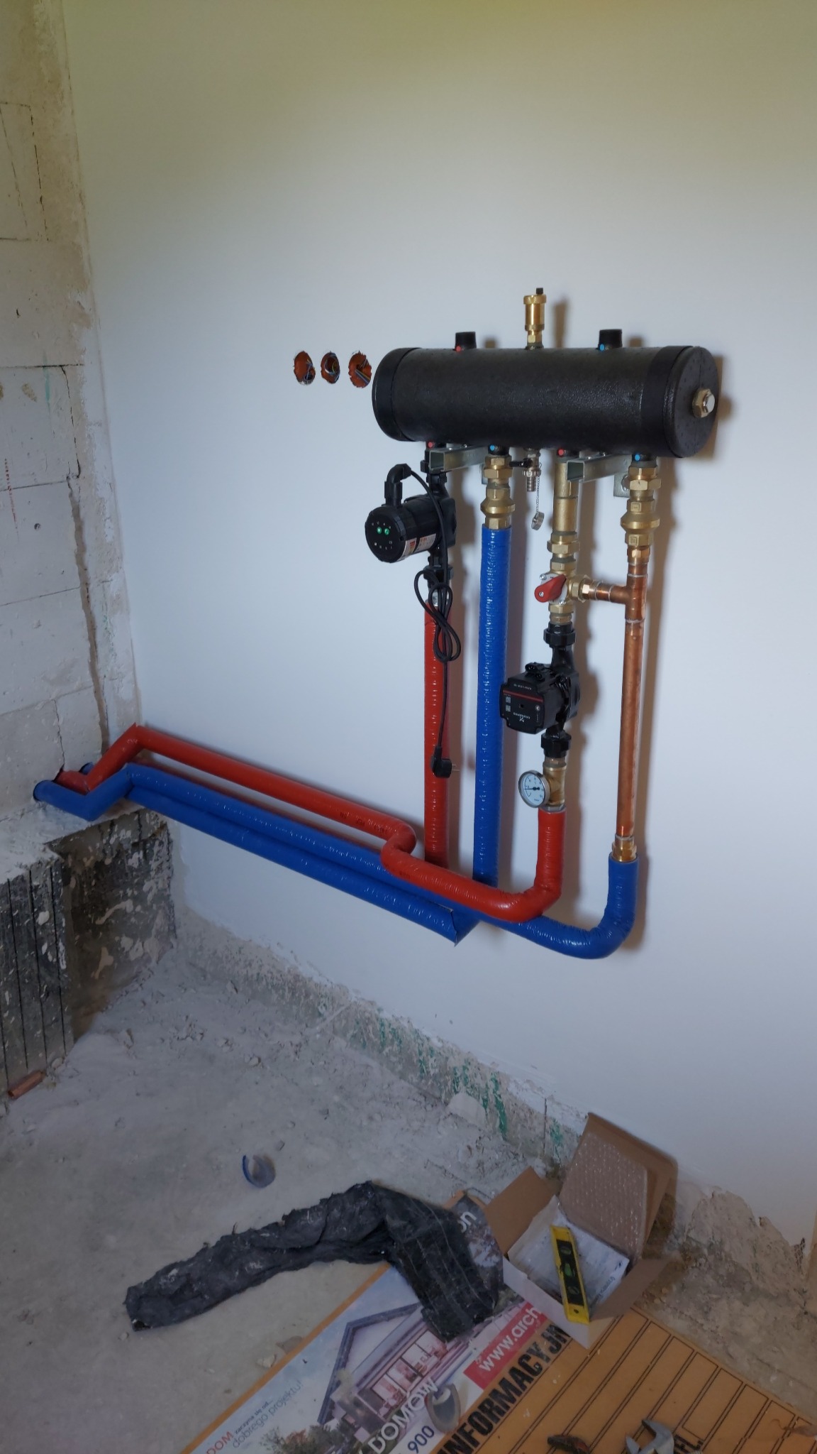 Rozdzielacz centralnego ogrzewania z dwoma pompami obiegowymi, rurami w izolacji czerwonej i niebieskiej oraz miedzianymi rurami, zamontowany na białej ścianie w surowym stanie.