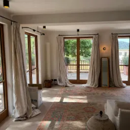 Przestronne wnętrze z dużymi oknami drewnianymi i zwisającymi zasłonami w odcieniach beżu, na podłodze wzorzysty dywan, widoczne elementy wyposażenia w trakcie przeprowadzki lub remontu, w tle...