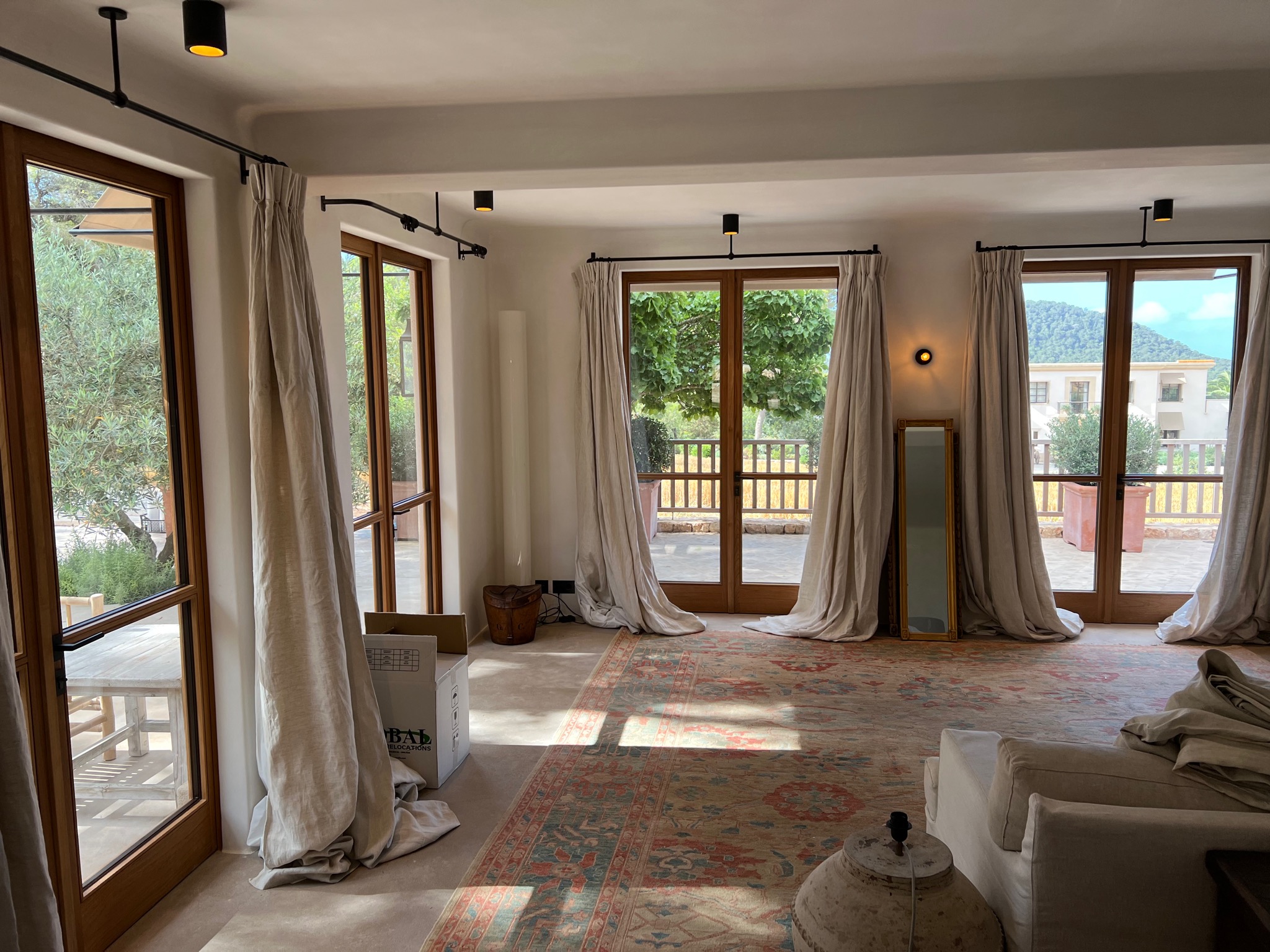 Przestronne wnętrze z dużymi oknami drewnianymi i zwisającymi zasłonami w odcieniach beżu, na podłodze wzorzysty dywan, widoczne elementy wyposażenia w trakcie przeprowadzki lub remontu, w tle...