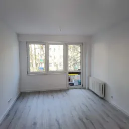 Pusty pokój po remoncie: świeżo pomalowane białe ściany, szare panele podłogowe imitujące drewno, biały grzejnik pod oknem, z wystającymi kablami ze stropu.