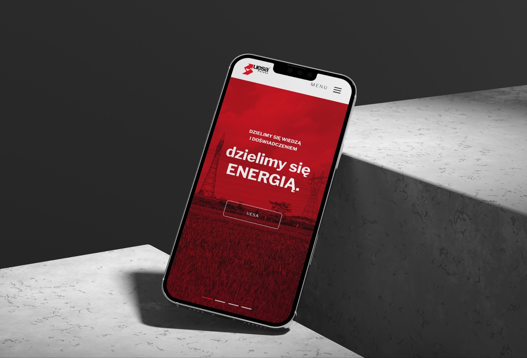 Smartfon wyświetla stronę internetową z czerwonym tłem, sloganem 'dzielimy się ENERGIĄ' i grafiką słupów wysokiego napięcia, umieszczony na szarej, teksturowanej powierzchni.