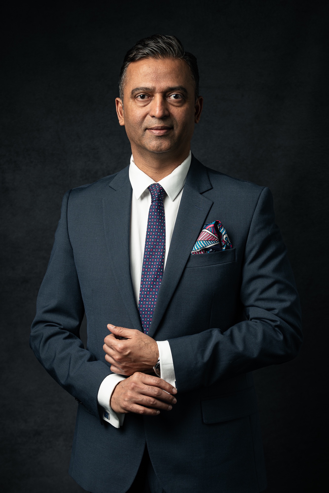 Portret biznesowy mężczyzny w granatowym garniturze, białej koszuli i wzorzystym krawacie, z kolorową poszetką w kieszonce, na ciemnoszarym tle.