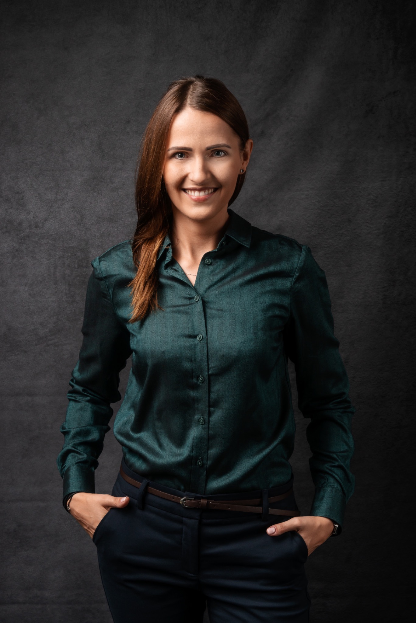 Portret uśmiechniętej kobiety w szmaragdowej koszuli i granatowych spodniach, ręce w kieszeniach, na ciemnym tle.