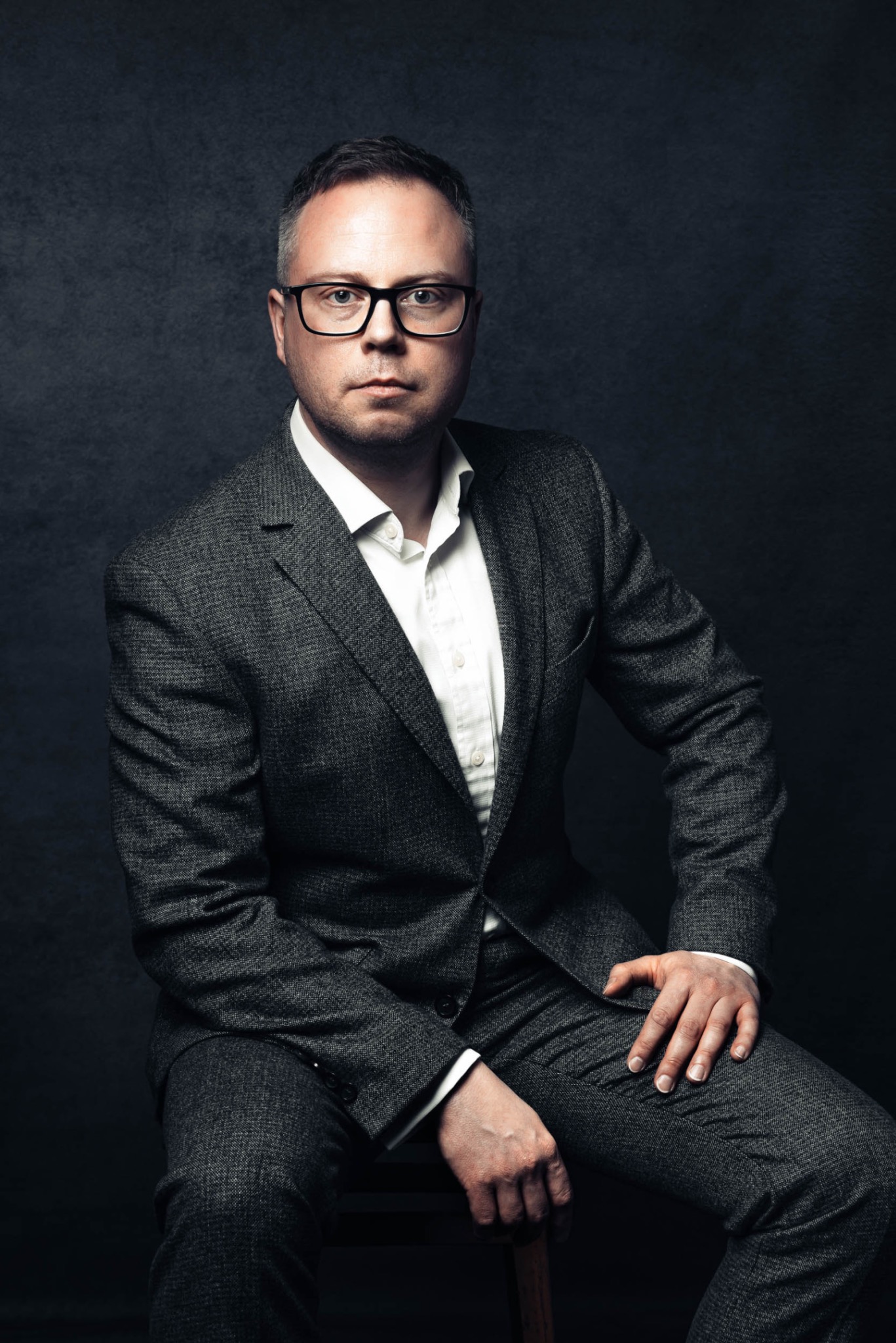 Mężczyzna w okularach i tweedowym garniturze, siedzący na drewnianym stołku na tle ciemnego, teksturowanego tła. Portret biznesowy z poważnym wyrazem twarzy.