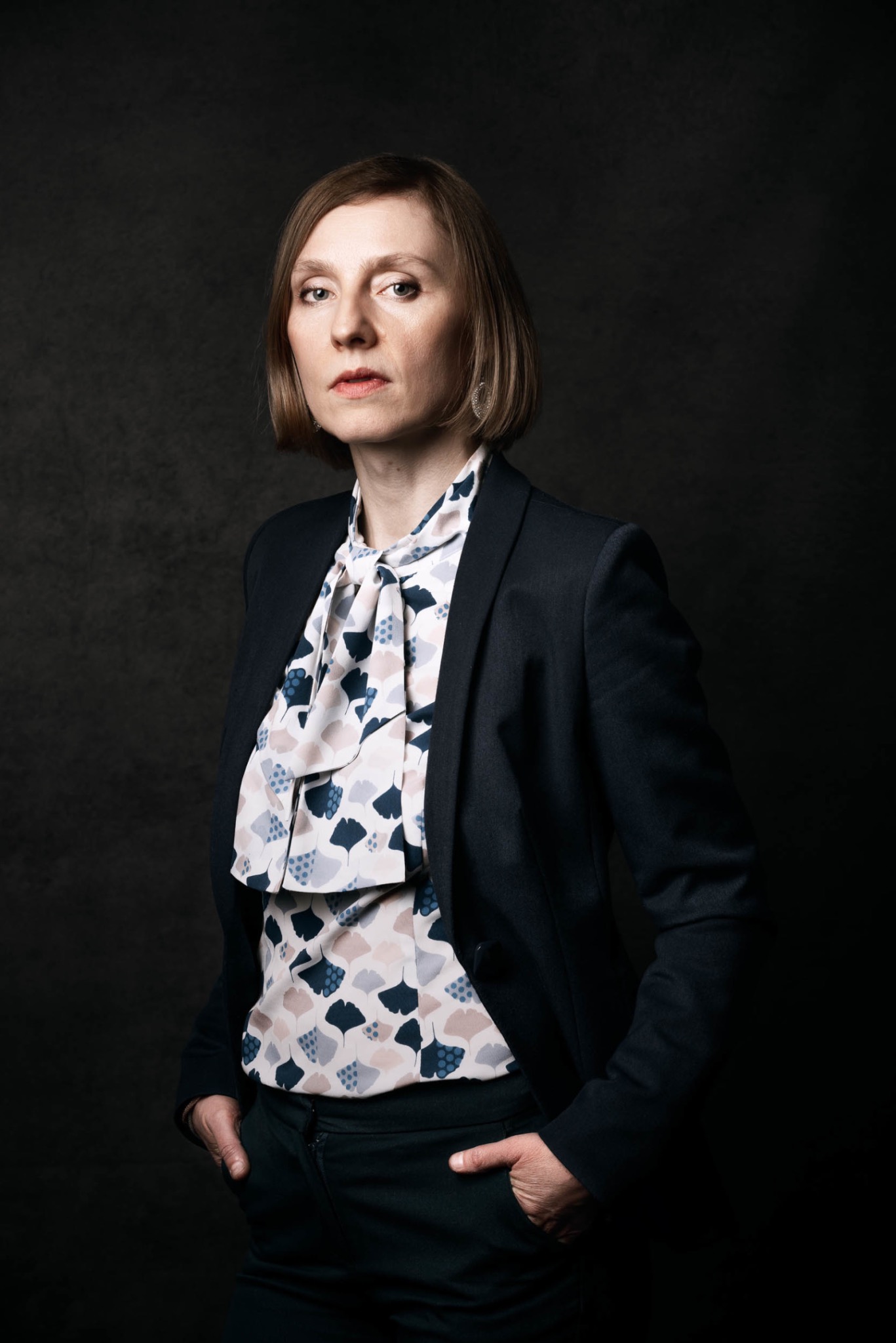 Portret kobiety w ciemnym garniturze i wzorzystej bluzce z kokardą, ręce w kieszeniach, na ciemnym tle.