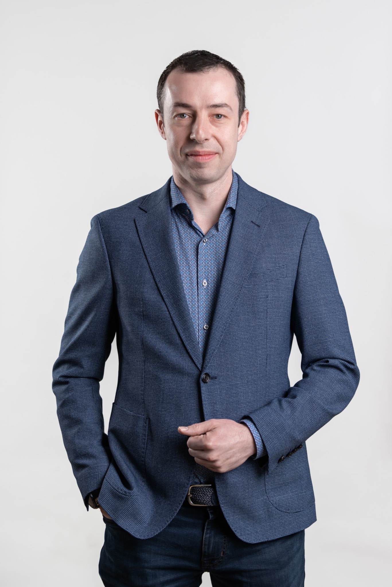 Portret mężczyzny w niebieskiej marynarce i wzorzystej koszuli, pozującego na jasnym tle. Lewa ręka w kieszeni, prawa dłoń ułożona na marynarce.