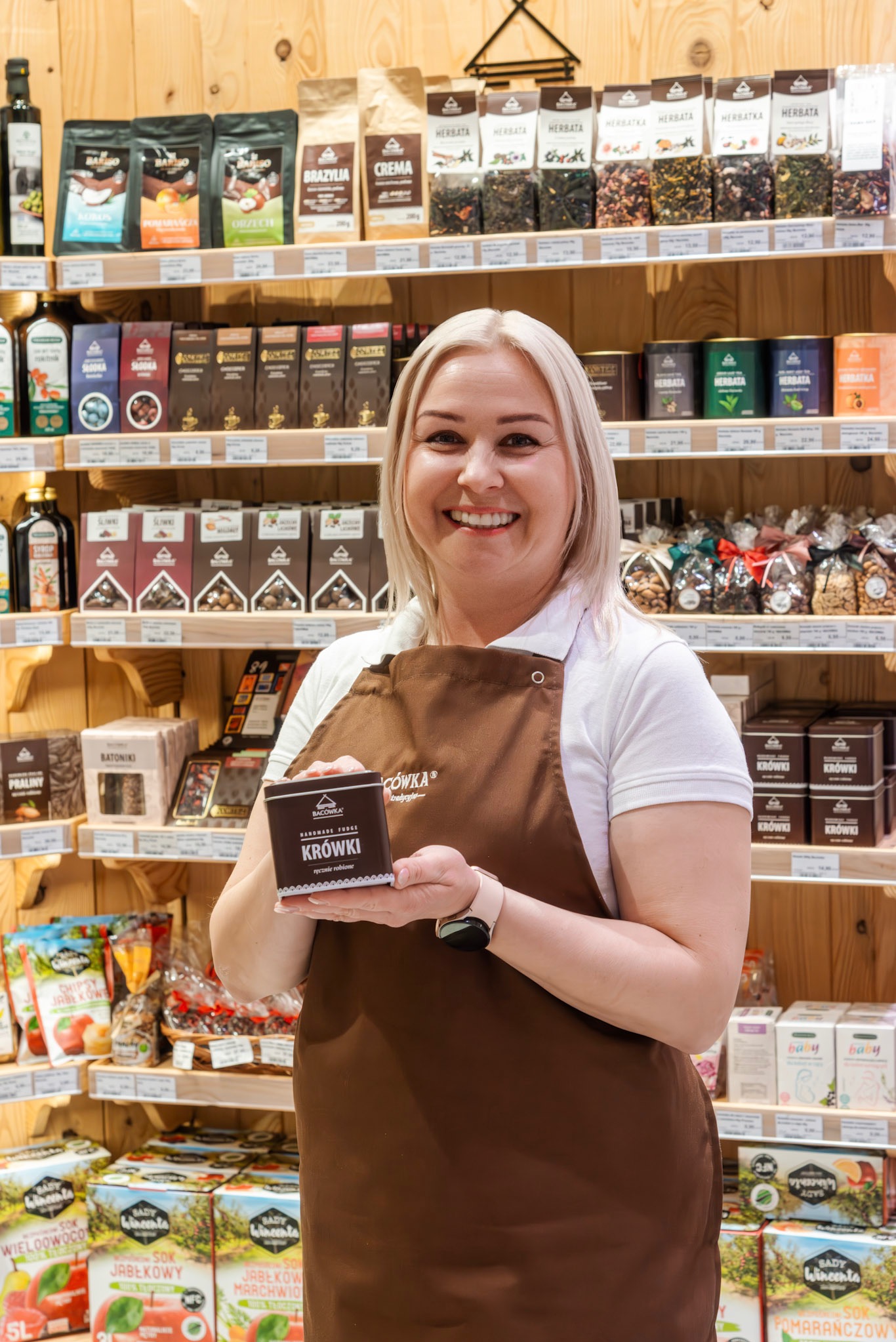 Uśmiechnięta blondynka w brązowym fartuchu prezentuje puszkę ręcznie robionych krówek Bacówka na tle półek z herbatami, kawami i słodyczami w sklepie.