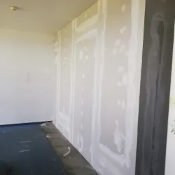 Ściana z płyt kartonowo-gipsowych w trakcie szpachlowania, widoczne łączenia płyt i miejsca po wkrętach wypełnione masą szpachlową, sufit z perforowanymi panelami akustycznymi.