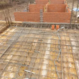 Budowa domu jednorodzinnego: widok z góry na zbrojenie płyty fundamentowej i częściowo wzniesione ściany z czerwonej cegły, widoczne pomarańczowe rury wentylacyjne.