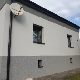 Dwukolorowa elewacja domu jednorodzinnego z widocznym fragmentem instalacji satelitarnej i pracującym obok pracownikiem, tynk strukturalny na dolnej części ściany.