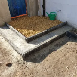 Ułożone schody z kostki brukowej prowadzące do drzwi zabezpieczonych folią, obok zielony konewka i wysypany kamień ozdobny.