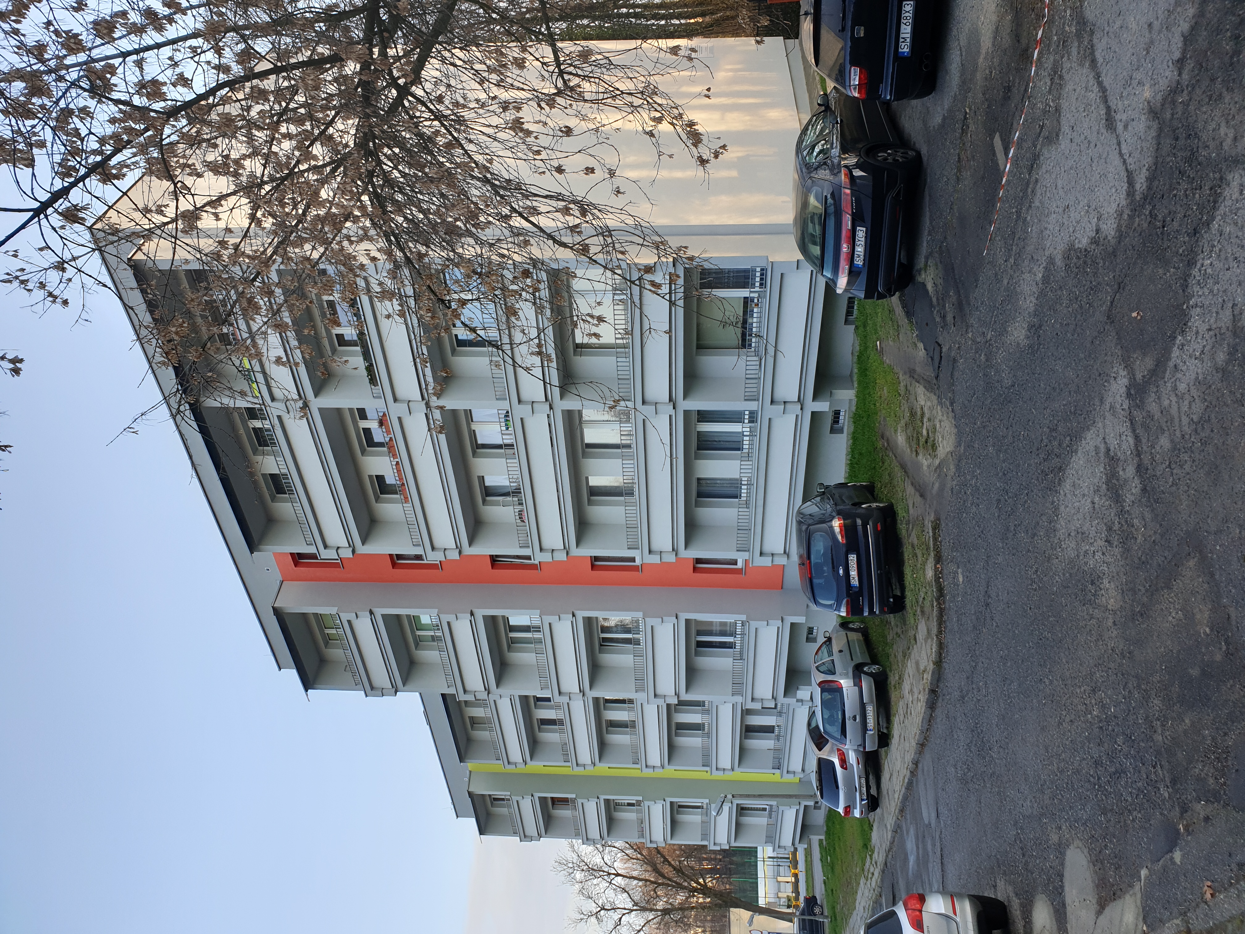 Wielopiętrowy blok mieszkalny z balkonami, częściowo ocieplony i pomalowany na szaro, z pomarańczowym pasem na górze i zielonym na dole, widoczny z poziomu ulicy z zaparkowanymi samochodami...