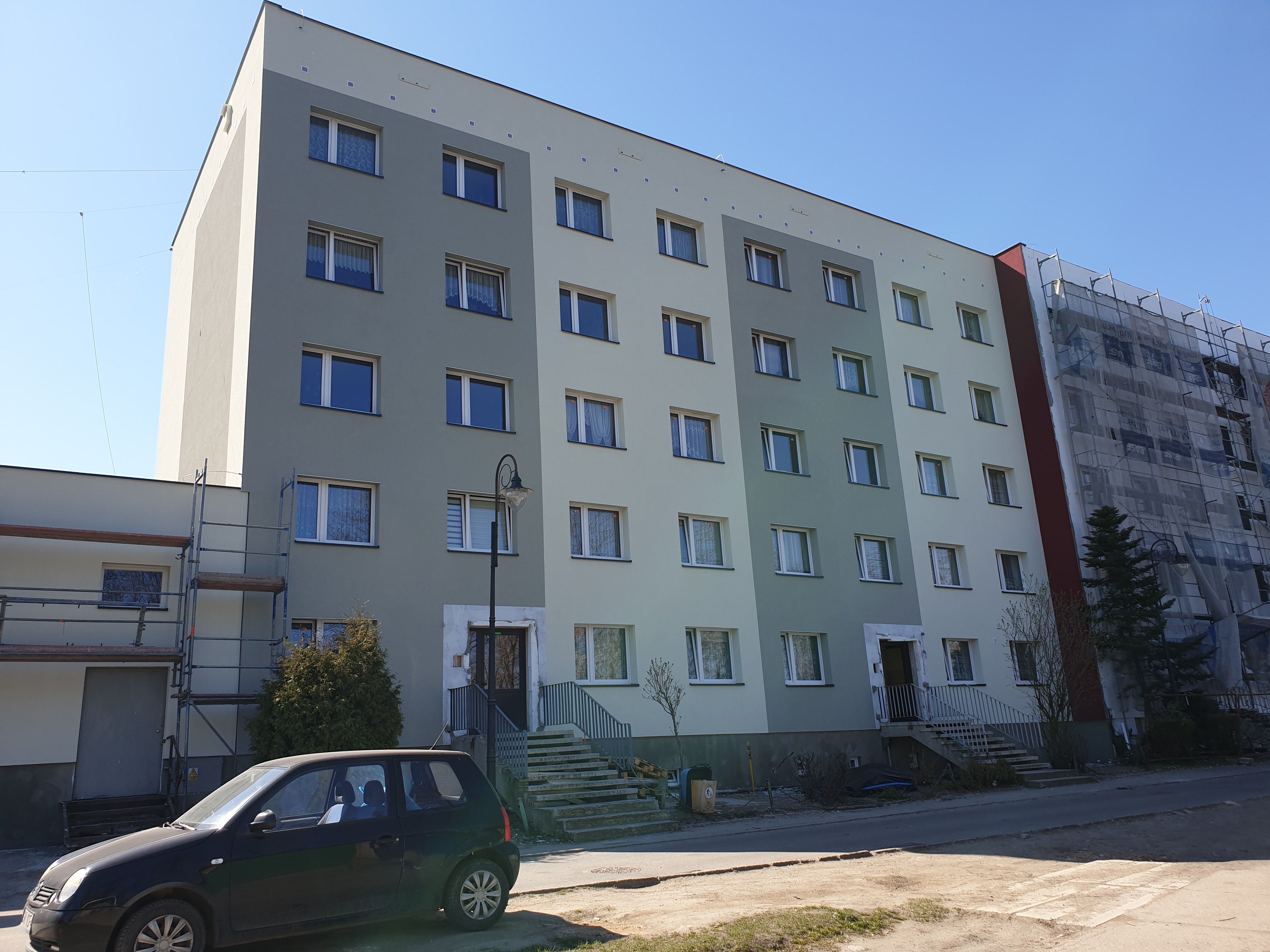 Wielopiętrowy budynek mieszkalny po renowacji elewacji, z fragmentem elewacji w trakcie prac pod rusztowaniem, widoczny czarny samochód osobowy na pierwszym planie.
