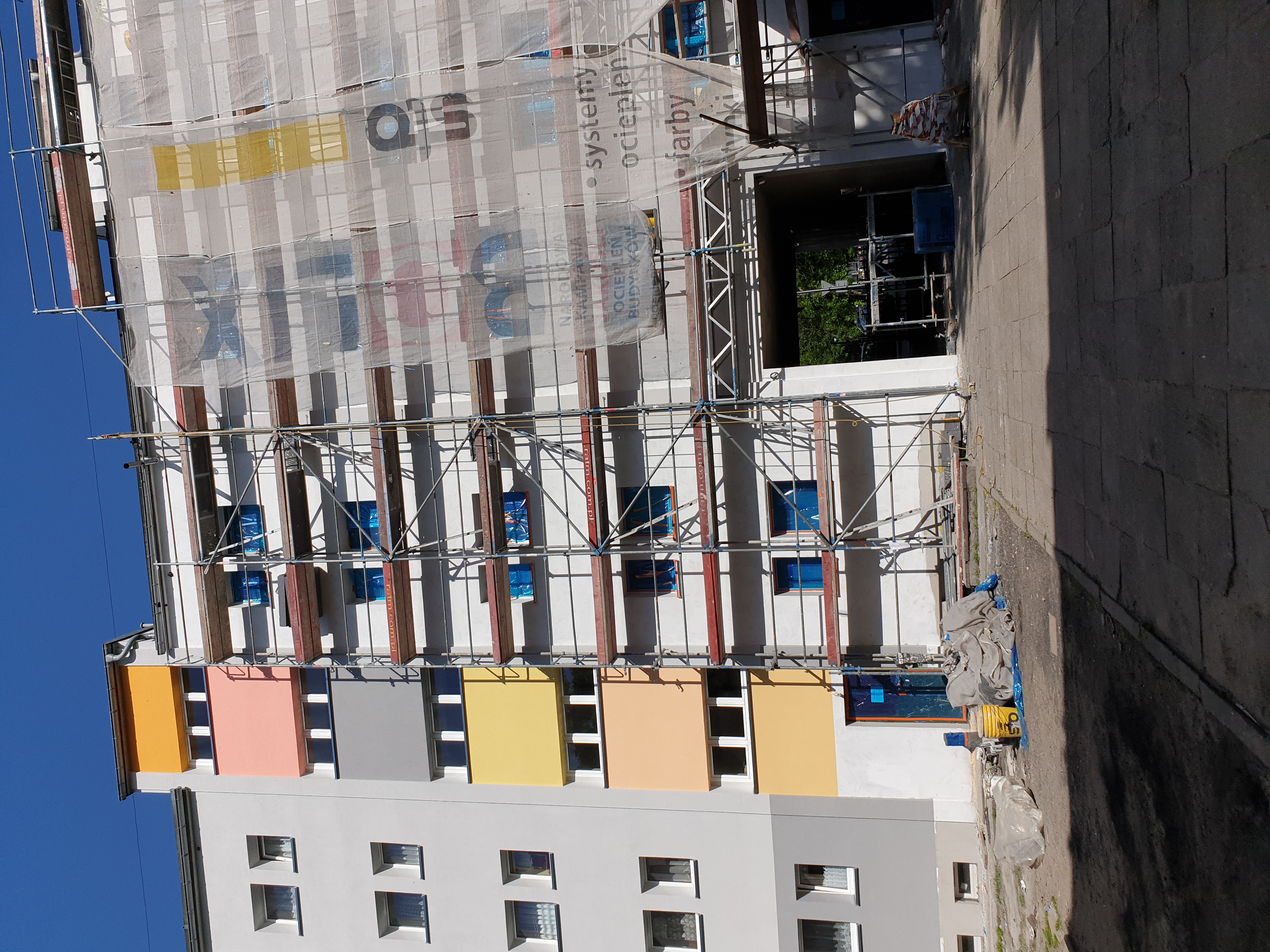 Wielopiętrowy budynek w trakcie prac elewacyjnych, z widocznymi rusztowaniami, siatką ochronną z nadrukiem reklamowym oraz fragmentami pomalowanej elewacji w pastelowych kolorach.