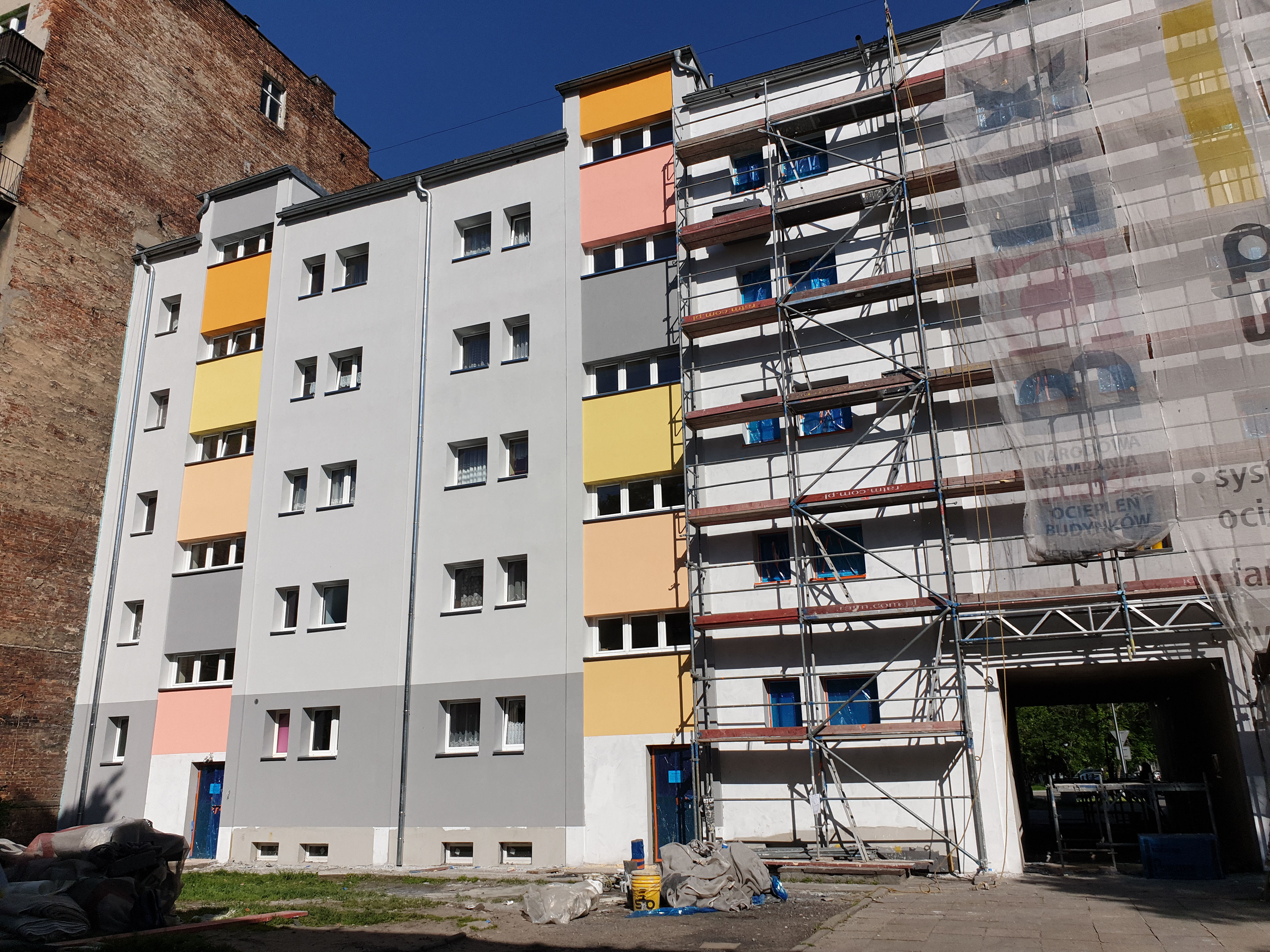 Wielopiętrowy budynek w trakcie ocieplania elewacji, widoczne rusztowania, zabezpieczone okna, kolorowe pasy na fasadzie oraz sterta worków z materiałami budowlanymi u podstawy budynku.