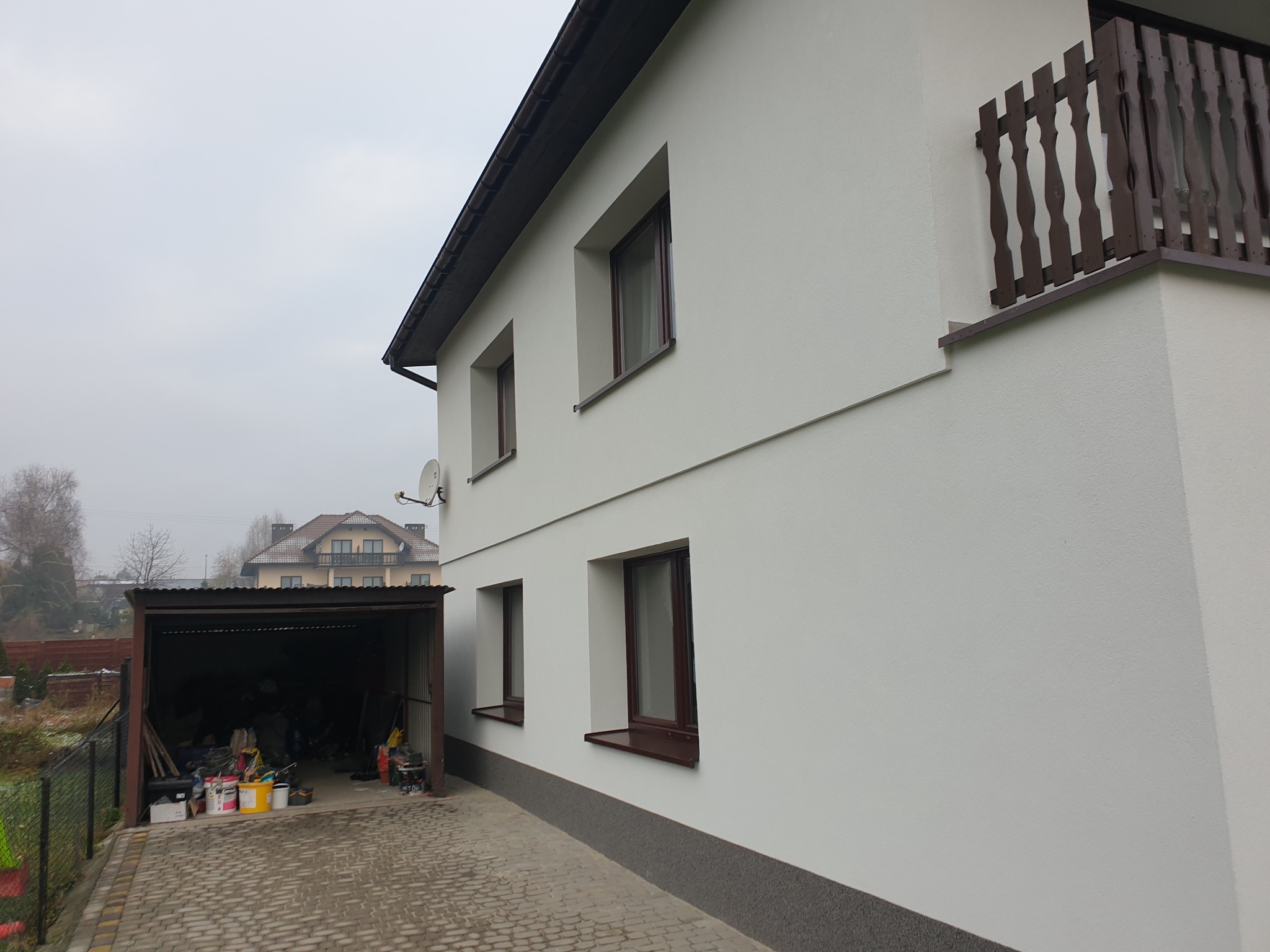 Biała elewacja domu jednorodzinnego z brązowymi oknami i balustradą balkonu, widoczny garaż z farbami i narzędziami, w tle inny dom i pochmurne niebo.