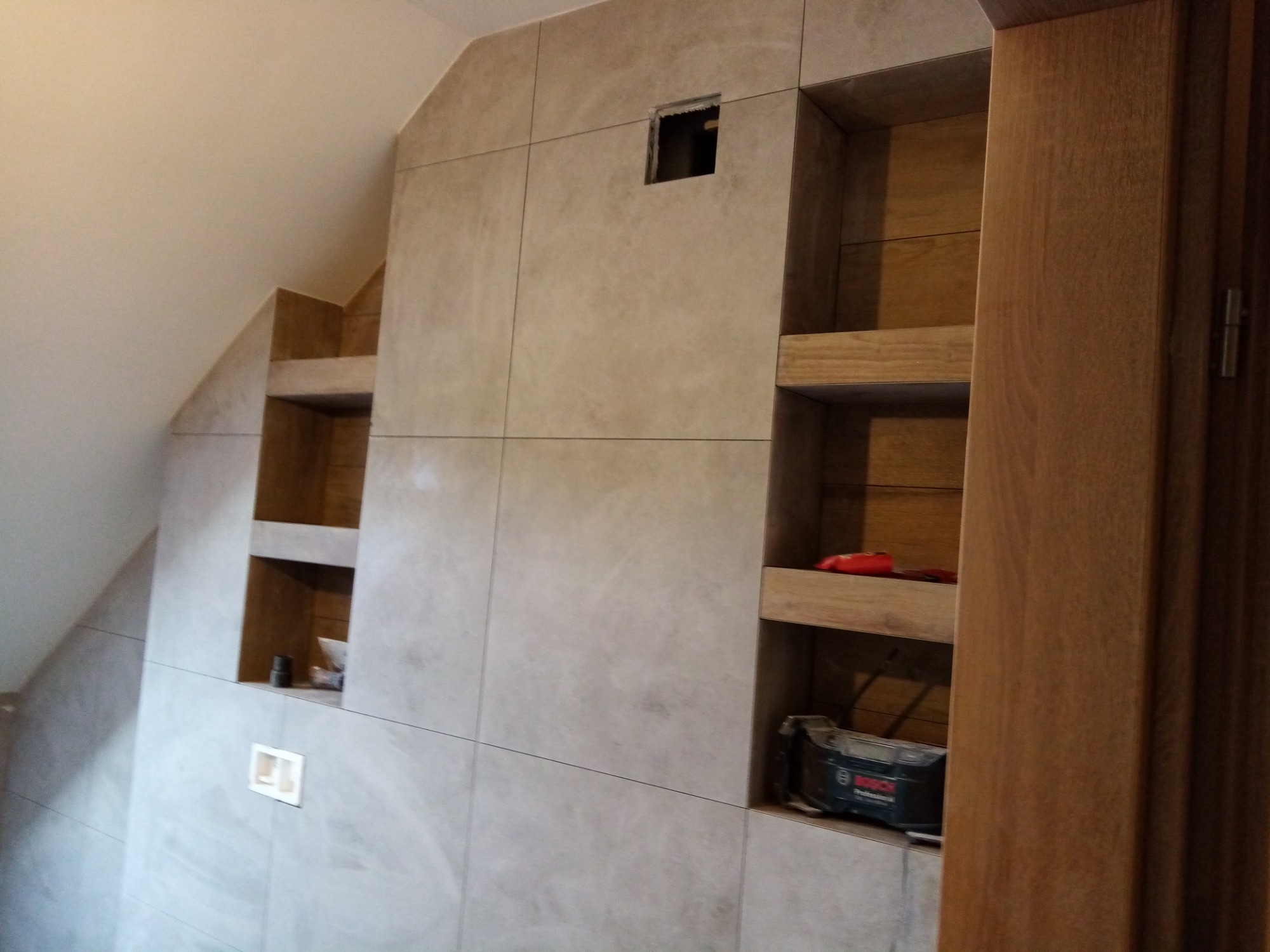 Ściana łazienki wykończona płytkami w odcieniach beżu z wnękami, w jednej z nich widoczny otwór instalacyjny, w innych narzędzia budowlane, fragment drewnianej ościeżnicy drzwiowej po prawej.