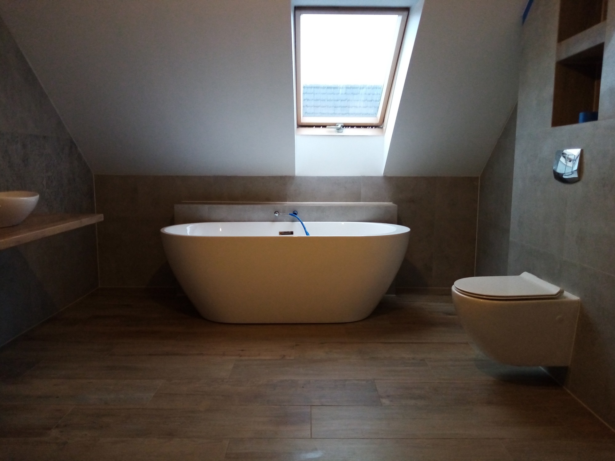 Nowoczesna łazienka na poddaszu z wolnostojącą wanną, podwieszaną toaletą i drewnopodobnymi płytkami podłogowymi, oświetlona przez okno dachowe.