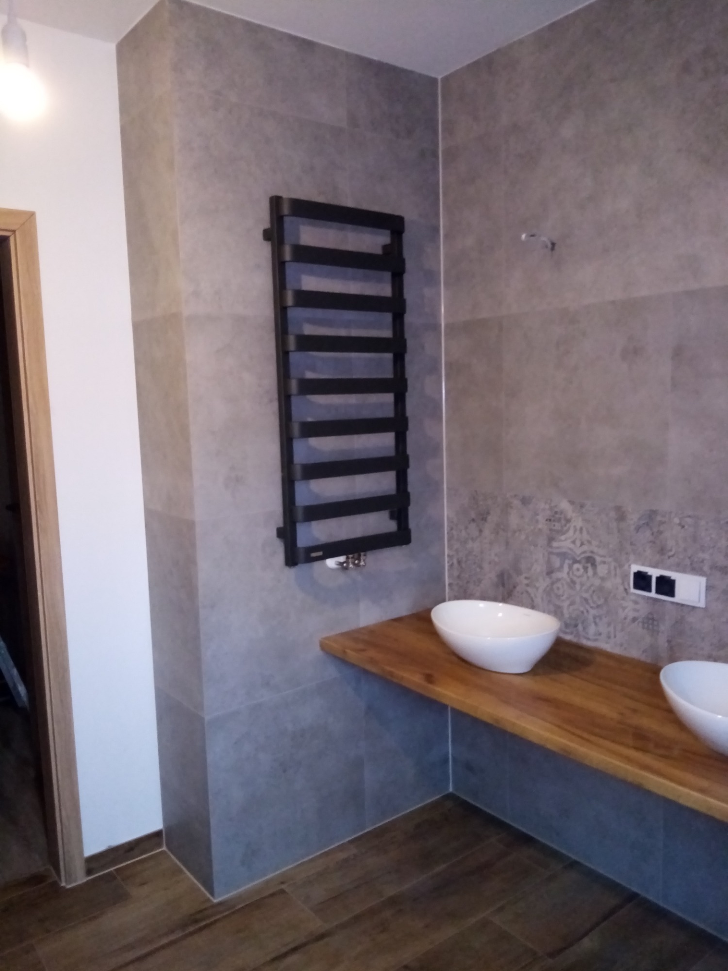 Nowoczesna łazienka z szarymi płytkami imitującymi kamień, drewnianym blatem pod umywalki nablatowe i czarnym grzejnikiem drabinkowym.