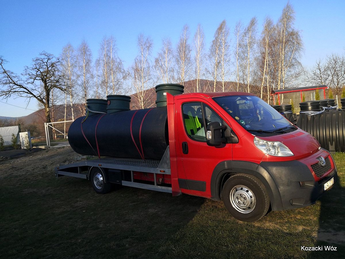 Czerwony samochód dostawczy z platformą transportową przewożący zbiornik na szambo z trzema kominami, na tle drzew i gór.