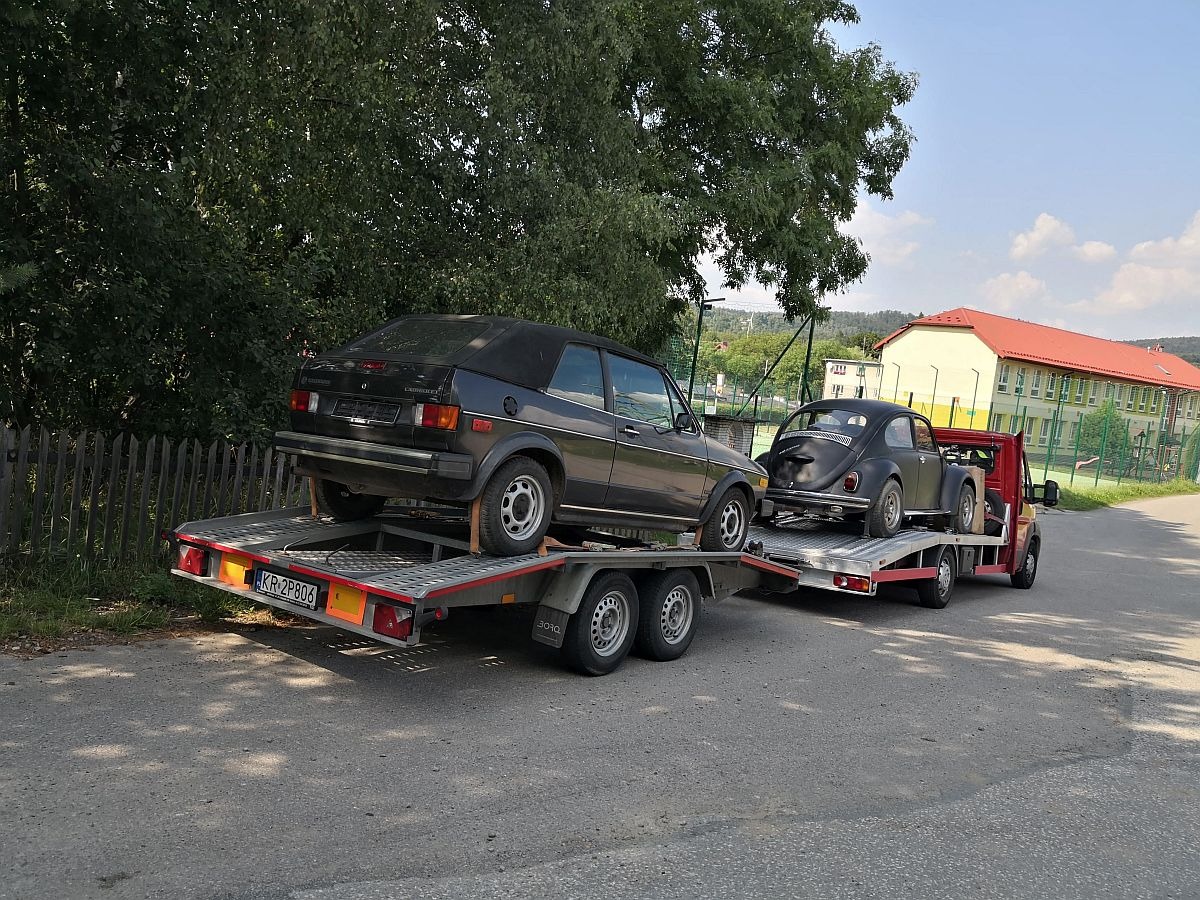 Dwa czarne samochody, Volkswagen Garbus i kabriolet, przewożone na przyczepach transportowych, z budynkiem szkolnym z czerwonym dachem w tle.