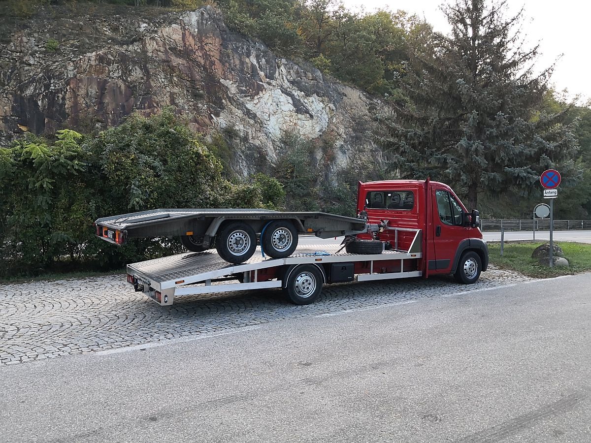 Czerwony samochód z platformą transportową na przyczepę, zaparkowany na kostce brukowej przy drodze, z górą i drzewami w tle.