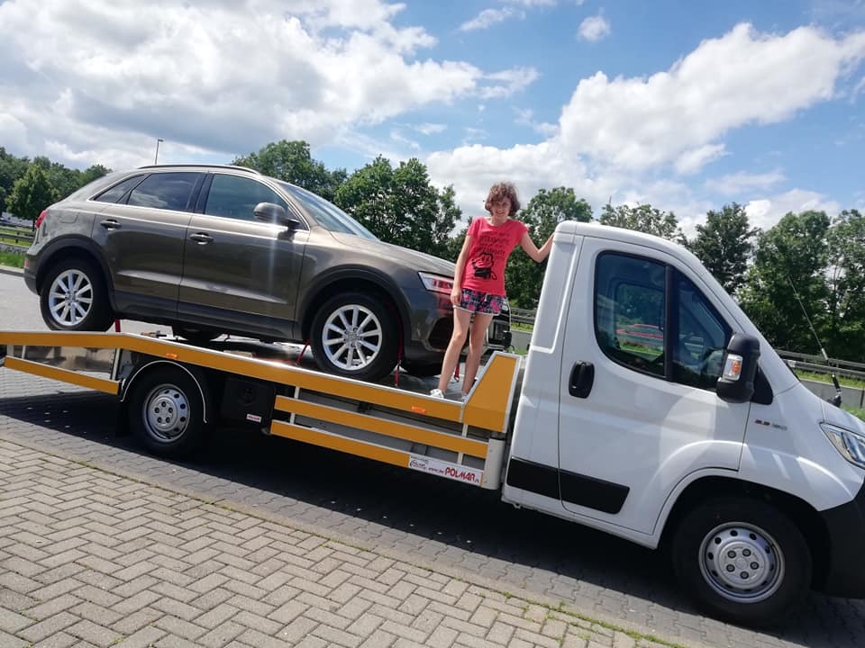 Brązowy SUV Audi Q3 transportowany na białej platformie pomocy drogowej z żółtymi elementami zabezpieczającymi; dziewczyna w różowej koszulce opiera się o kabinę pojazdu na tle pochmurnego nieba.