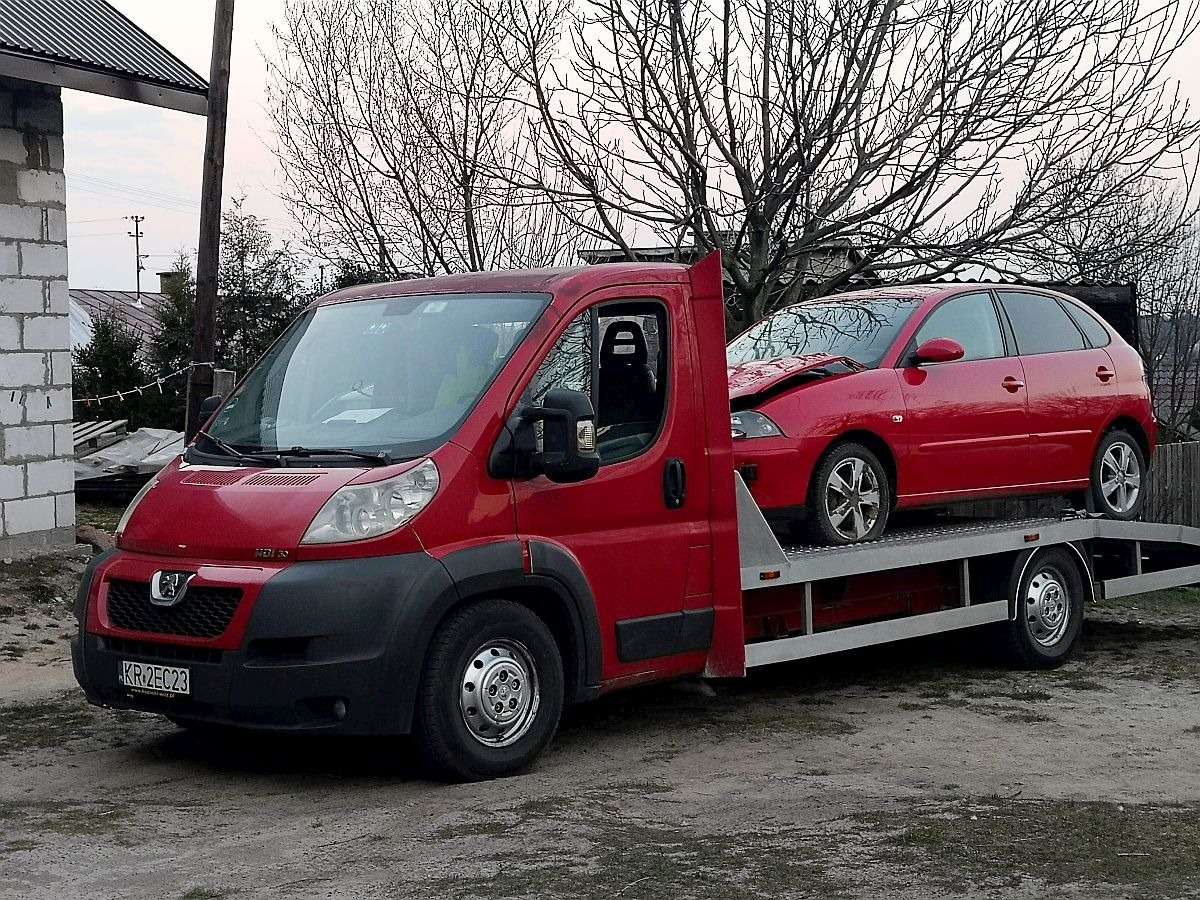 Czerwona platforma transportowa Peugeot z uszkodzonym czerwonym samochodem osobowym na platformie, zaparkowana na szutrowej drodze przed budynkiem w trakcie zachmurzonego dnia.