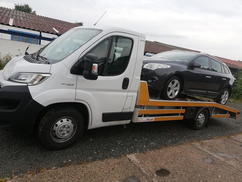 Biały samochód dostawczy z platformą transportową przewozi szary samochód osobowy na tle budynku z czerwoną dachówką.