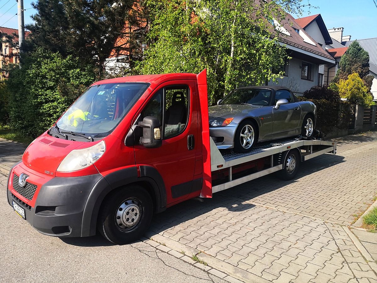 Czerwona laweta Fiat Ducato z załadowanym srebrnym sportowym kabrioletem Honda S2000 na tle domów jednorodzinnych i zieleni.