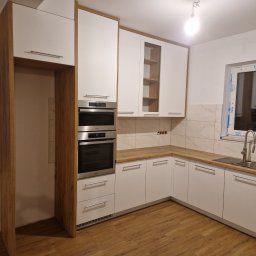 Firma SEMA - Nowoczesna kuchnia na wymiar z białymi frontami i drewnianymi akcentami. Widoczne zabudowane piekarniki i płyta grzewcza, drewniany blat i okno z widokiem na zewnątrz.