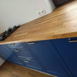 Firma SEMA - Granatowa zabudowa kuchenna z drewnianym blatem i złotymi uchwytami, widok z perspektywy podłogi, płyta gazowa wkomponowana w blat.