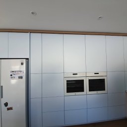 Firma SEMA - Biała zabudowa kuchenna z dwoma piekarnikami i lodówką Samsung, wykończona drewnianą ramą. Na lodówce rysunki i magnesy. Minimalistyczny design.