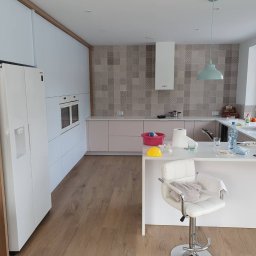 Firma SEMA - Jasna kuchnia z białymi frontami i drewnianymi akcentami. Widoczny blat z wyspą, krzesło barowe i sprzęt AGD. Na blacie naczynia i akcesoria kuchenne.