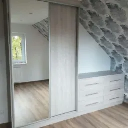 Jasna szafa wnękowa z przesuwnymi drzwiami lustrzanymi, wkomponowana w skos pomieszczenia, z komodą z szufladami po prawej stronie, na tle ściany w pióra.