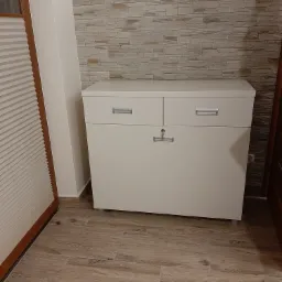 Biała komoda z dwoma szufladami i zamykaną szafką na tle ściany wyłożonej kamieniem dekoracyjnym, widoczna na drewnopodobnej podłodze.