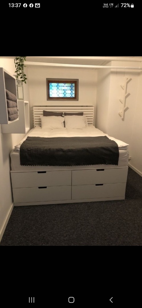 Białe łóżko z szufladami w minimalistycznej sypialni. Nad łóżkiem okno z witrażem i półki na ręczniki. Dekoracyjny wieszak na ścianie. Ciemna wykładzina podłogowa.