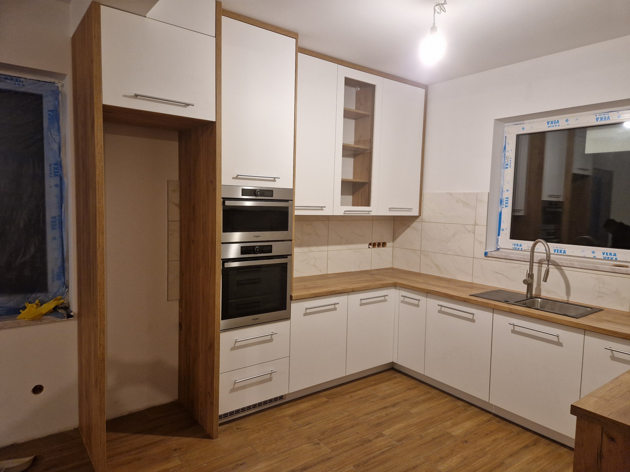 Nowoczesna kuchnia na wymiar z białymi frontami i drewnianymi akcentami. Widoczne zabudowane piekarniki i płyta grzewcza, drewniany blat i okno z widokiem na zewnątrz.