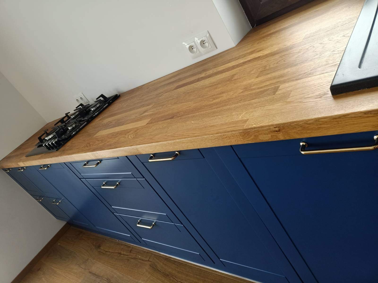 Granatowa zabudowa kuchenna z drewnianym blatem i złotymi uchwytami, widok z perspektywy podłogi, płyta gazowa wkomponowana w blat.