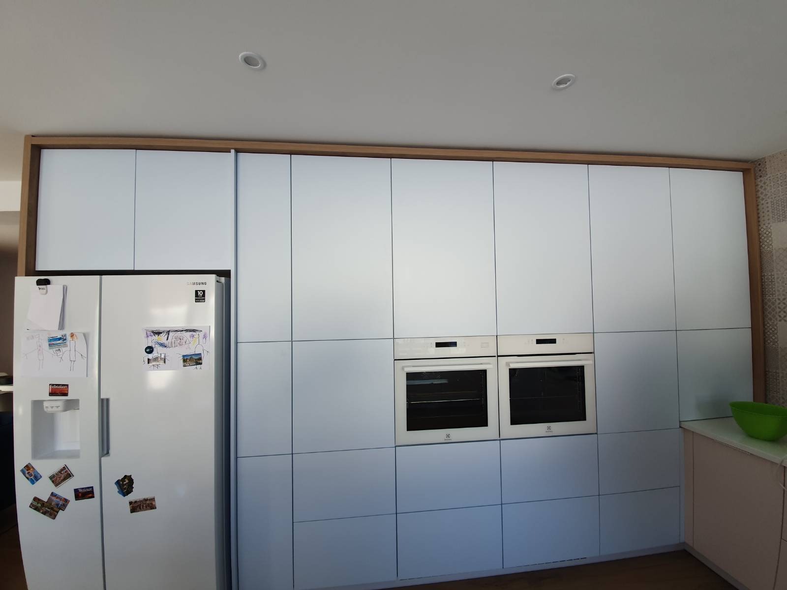 Biała zabudowa kuchenna z dwoma piekarnikami i lodówką Samsung, wykończona drewnianą ramą. Na lodówce rysunki i magnesy. Minimalistyczny design.